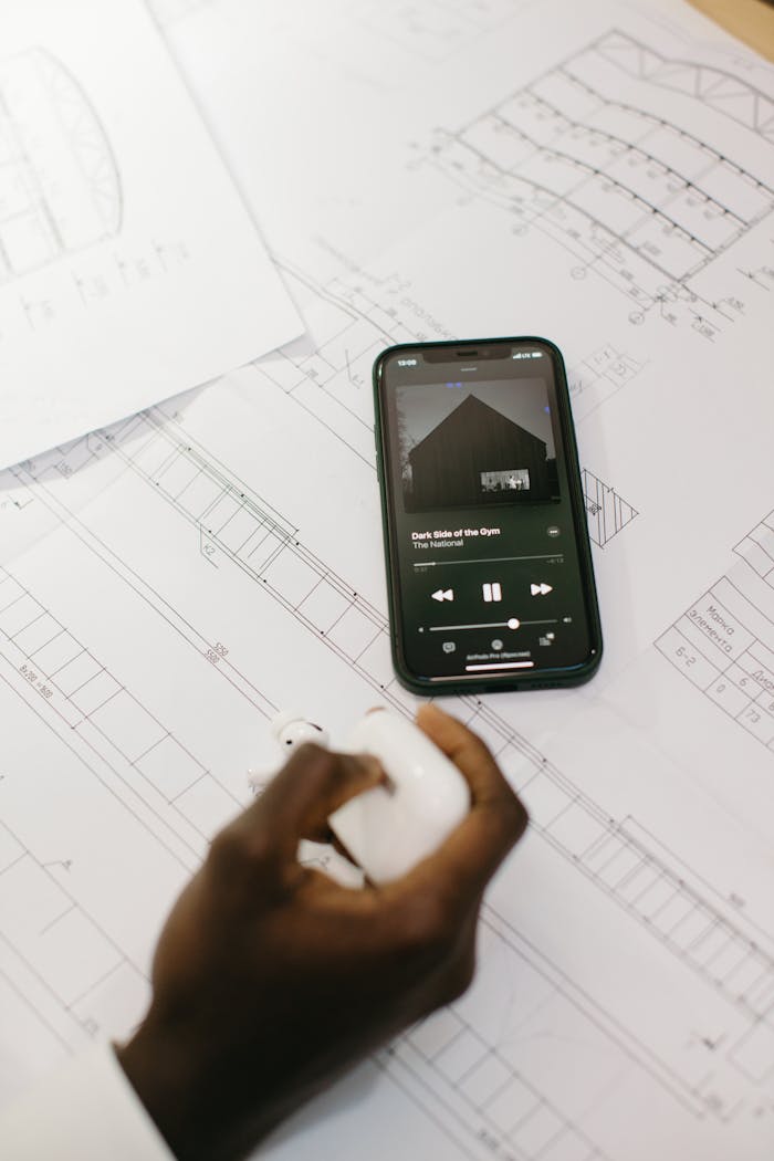 A smartphone playing music on architectural blueprints with a hand holding earbuds.