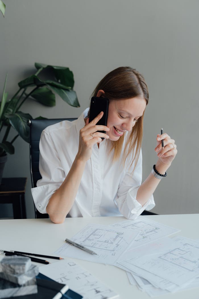 Businesswoman smiling while on a phone call, reviewing architectural plans in a modern office setting.